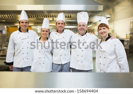 Five happy chefs smiling at the camera in a kitchen wearing uniforms