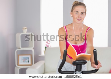 Sporty cheerful blonde training on exercise bike reading a book in bright living room