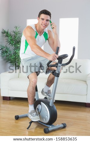 Cheerful handsome man training on exercise bike phoning in bright living room