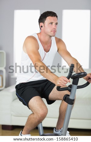 Handsome sporty man exercising on bike and listening to music in bright living room