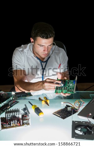 Attractive computer engineer examining hardware with stethoscope by night at his lit desk