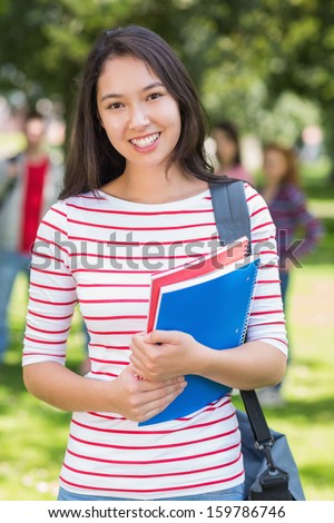 Portrait of college girl holding books with blurred students standing in the park