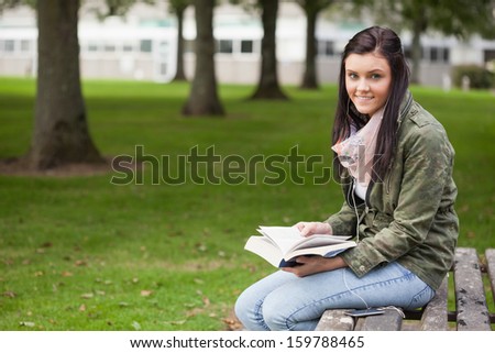 Cheerful brunette student sitting on bench reading on campus at college