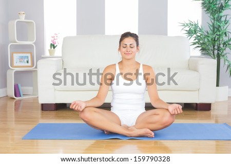 Front view of fit brunette woman relaxing in lotus position on an exercise mat in her living room