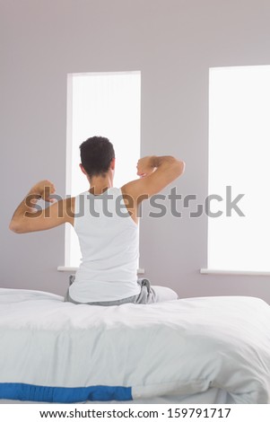 Rear view of man in pajamas sitting on bed stretching his arms in bright bedroom