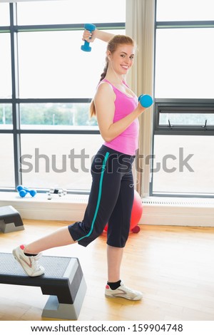 Beautiful young woman performing step aerobics exercise with dumbbells in a gym