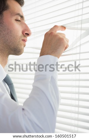 Attractive stern businessman spying through roller blind in bright office