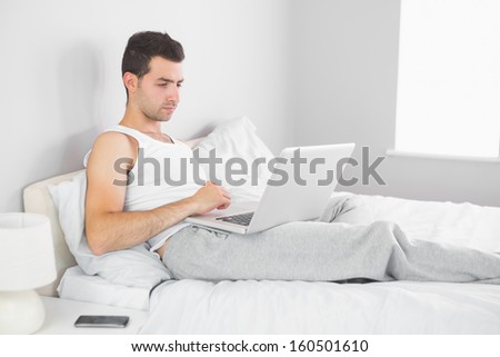 Handsome calm man using laptop in his bed in bright bedroom