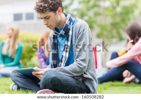 Serious young college boy using tablet PC with blurred students sitting in the park