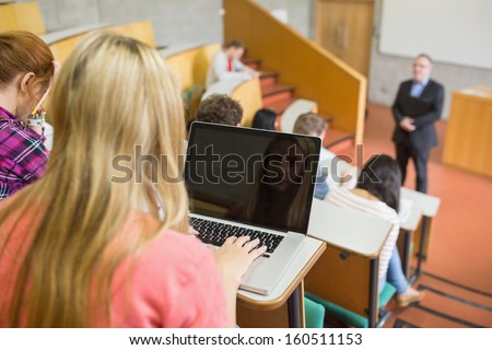 Rear view of a female using laptop with students and teacher at the college lecture hall