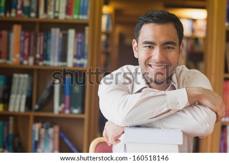 Cheerful attractive man posing leaning on a stack of books in library smiling at camera