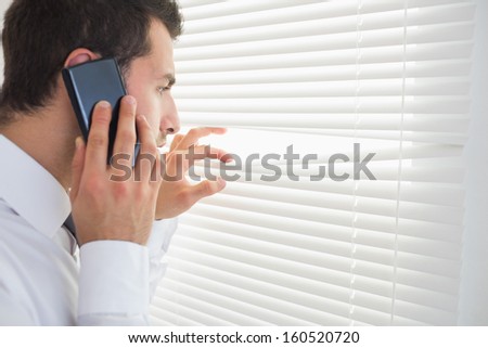 Serious businessman spying through roller blind while phoning in bright office