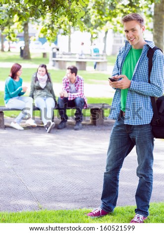 Portrait of a smiling college boy text messaging with blurred students sitting in the park