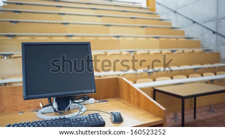 View of computer monitor with empty wooden seats with tables in a lecture hall