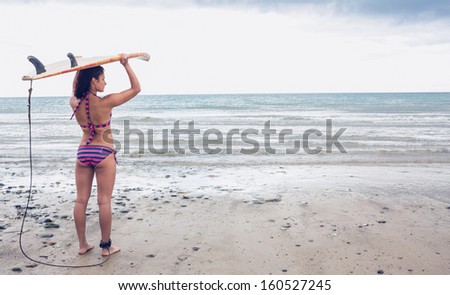 Rear view of a young bikini woman carrying surfboard on head at beach