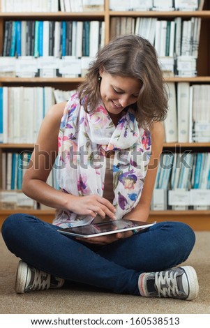 Full length of a happy female student against bookshelf using tablet PC on the library floor