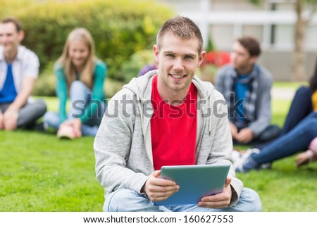 Portrait of a smiling college boy holding tablet PC with blurred students sitting in the park