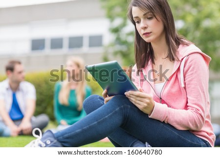 Serious young college girl using tablet PC with blurred students sitting in the park