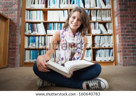 Full length of a happy female student sitting against bookshelf with a book on the library floor