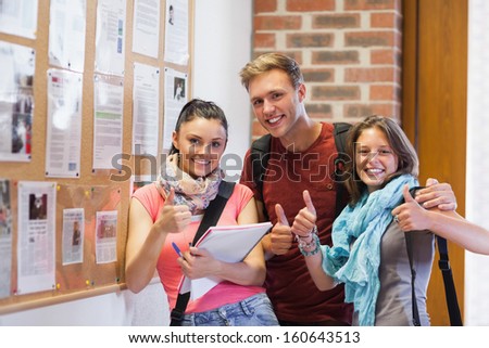 Three smiling students standing next to notice board showing thumbs up in school