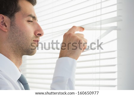 Attractive frowning businessman spying through roller blind in bright office