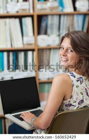 Portrait of a smiling female student against bookshelf using laptop in the library
