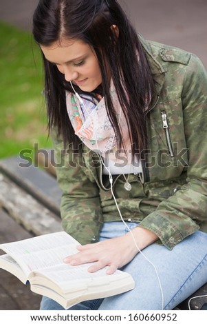 Content brunette student sitting on bench reading on campus at college