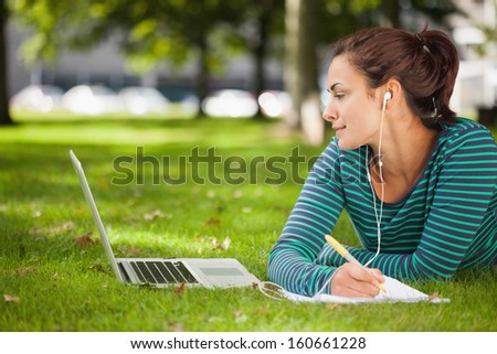 Attractive casual student lying on grass taking notes on campus at college