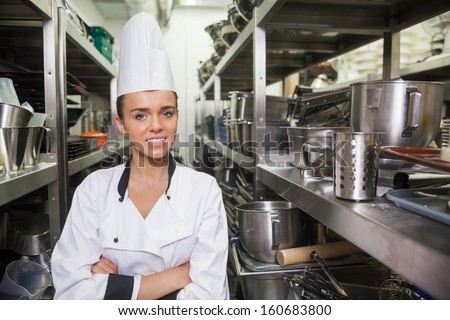Young cheerful chef standing arms crossed between shelves in professional kitchen