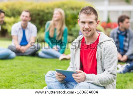 Portrait of a smiling college boy holding tablet PC with blurred students sitting in the park