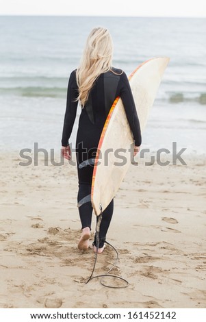 Full length rear view of a young woman in wet suit holding surfboard at beach