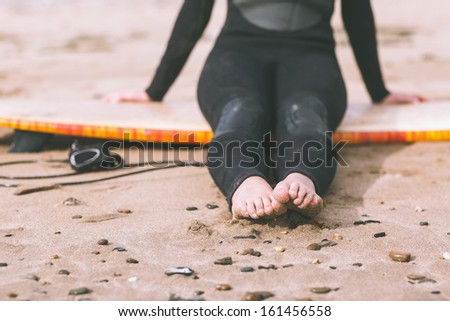 Low section of a young woman in wet suit with surfboard sitting at the beach