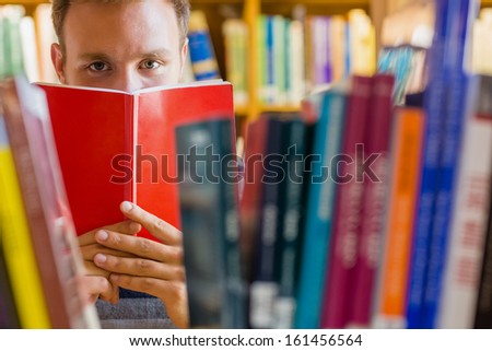 Close up of a young male student holding book in front of his face amid bookshelves in the college library