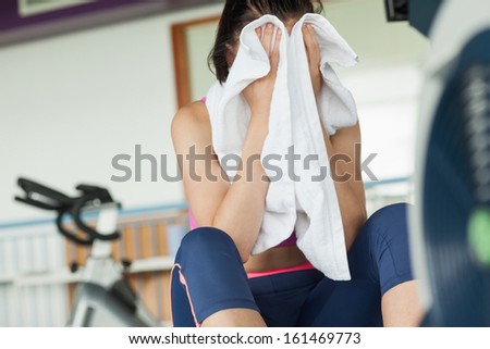 Tired young woman wiping face while working on row machine in fitness studio