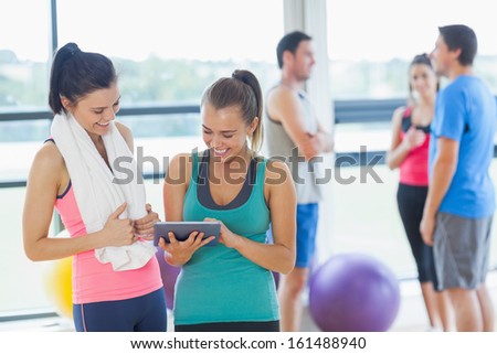 Fit women looking at digital table with friends chatting in background in bright exercise room
