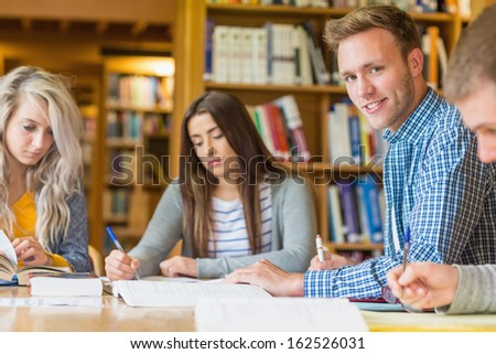 Portrait of a smiling male student with friends sitting at desk in the college library