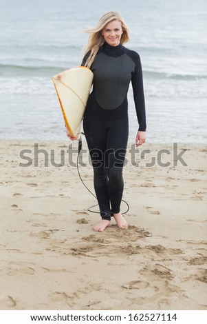 Full length portrait of a smiling young woman in wet suit holding surfboard at beach