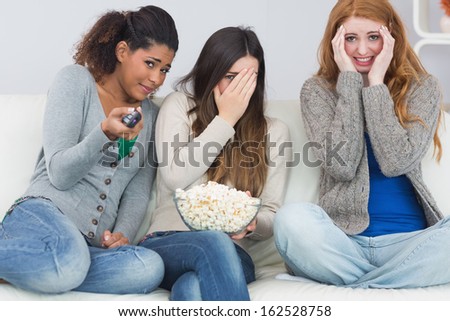 Portrait of scared young female friends with remote control and popcorn bowl on sofa at home