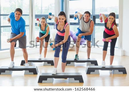 Full Length Of Instructor With Fitness Class Performing Step Aerobics Exercise With Dumbbells In A Gym