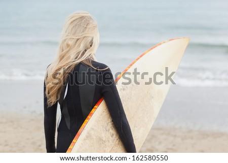 Rear view of a young woman in wet suit holding surfboard at beach
