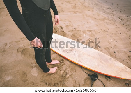 Low section of a young woman in wet suit with surfboard standing at the beach
