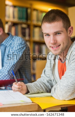 Portrait of a smiling male student with a friend sitting at desk in the college library