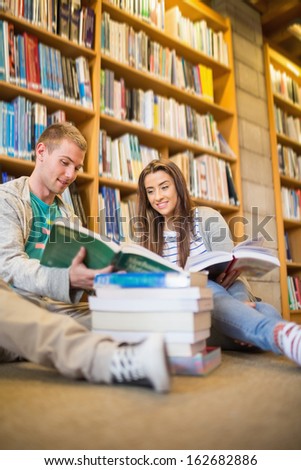 Two young students reading books against bookshelf while sitting on the library floor