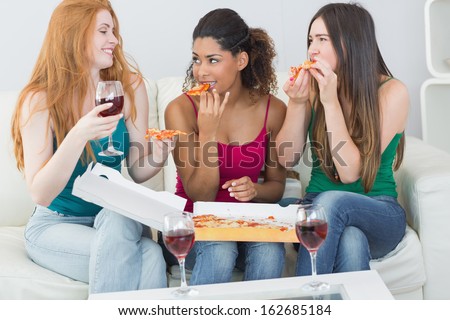 Happy young female friends eating pizza with wine on sofa at home
