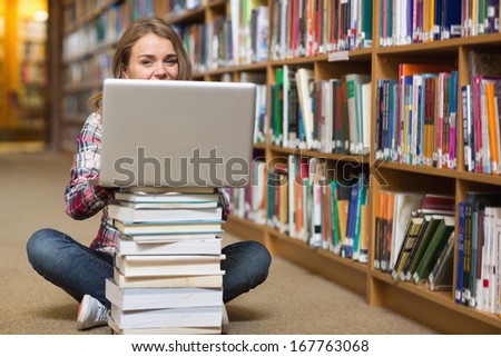 Happy student sitting on library floor using laptop on pile of books in college