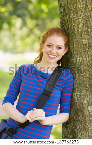Pretty redhead student leaning on tree looking at camera on college campus