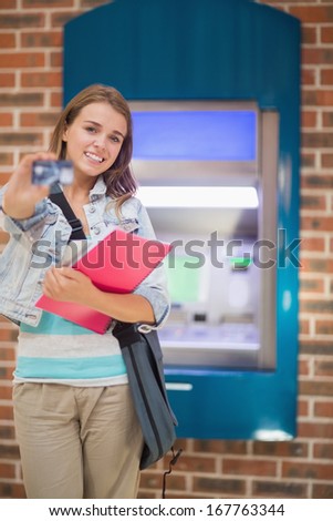 Happy student standing showing her card to camera at the atm in college