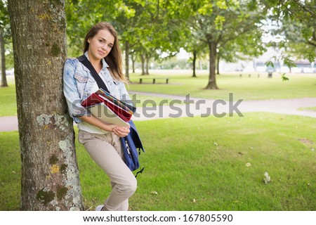 Cheerful student leaning on tree holding her books on college campus