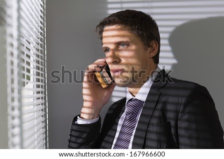 Serious young businessman peeking through blinds while on call in the office