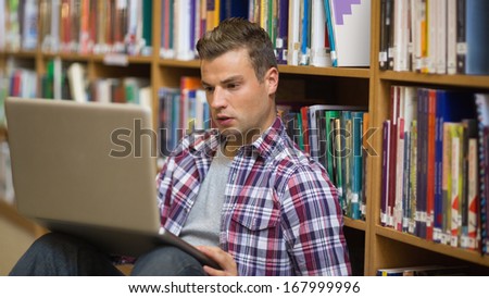 Serious young student sitting on library floor using laptop in college